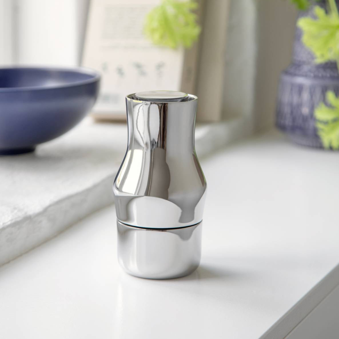 A silver Dorotea grinder made of polished SS304 stainless steel, featuring a ceramic CrushGrind mechanism, displayed on a white surface with a plant and bowl in the background.