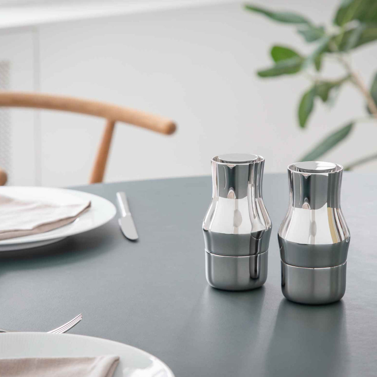 Two silver salt and pepper shakers on a table with plates and cutlery in the background. Gense Dorotea Collection Grinder German Design Award winner