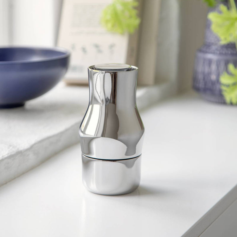 A silver Dorotea grinder made of polished SS304 stainless steel, featuring a ceramic CrushGrind mechanism, displayed on a white surface with a plant and bowl in the background.