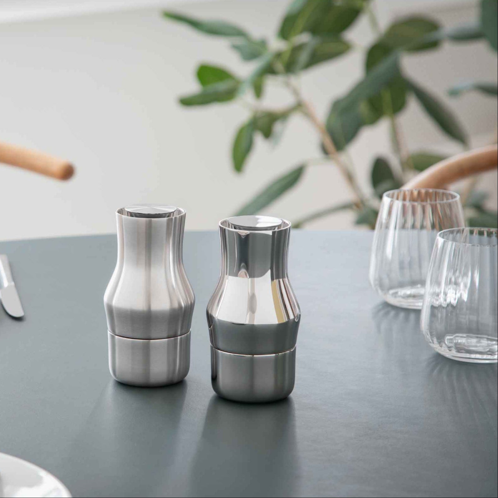 Two silver salt and pepper shakers on a table with glasses and a plant in the background. Gense Dorotea Collection Grinder German Design Award winner