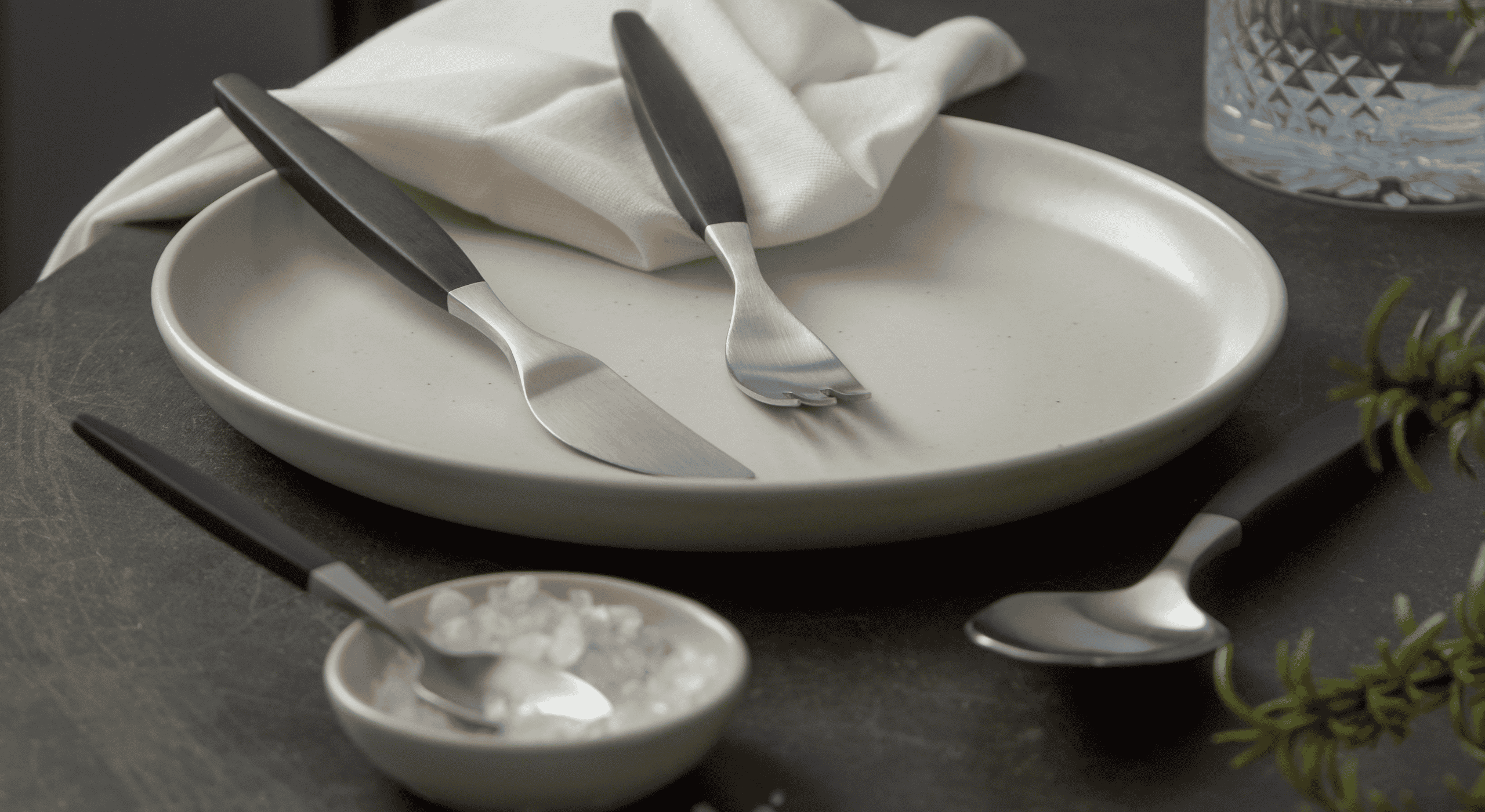Dining table setting with plates, cutlery, and a glass on a dark surface. Gense Focus collection