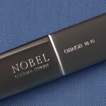 Close-up of a dark metallic cylindrical product labeled NOBEL and GEN5B 18-10, resting on a textured blue fabric background.
