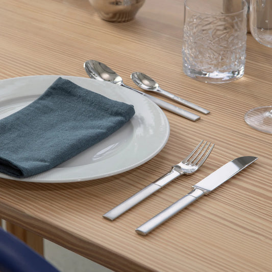 Dinner setting with a white plate, blue napkin, Gense Nobel Collection Cutlery, and glasses on a wooden table.