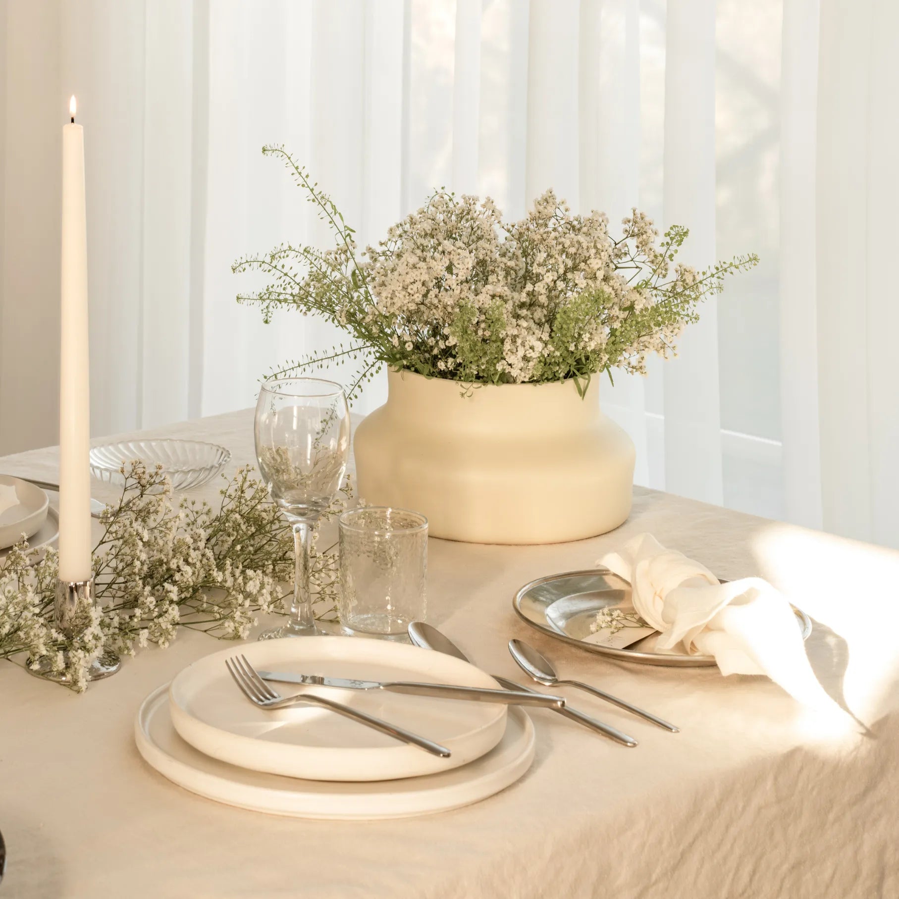 Elegant table setting with white plates, Gense Still Collection cutlery, and a vase of flowers on a light surface.