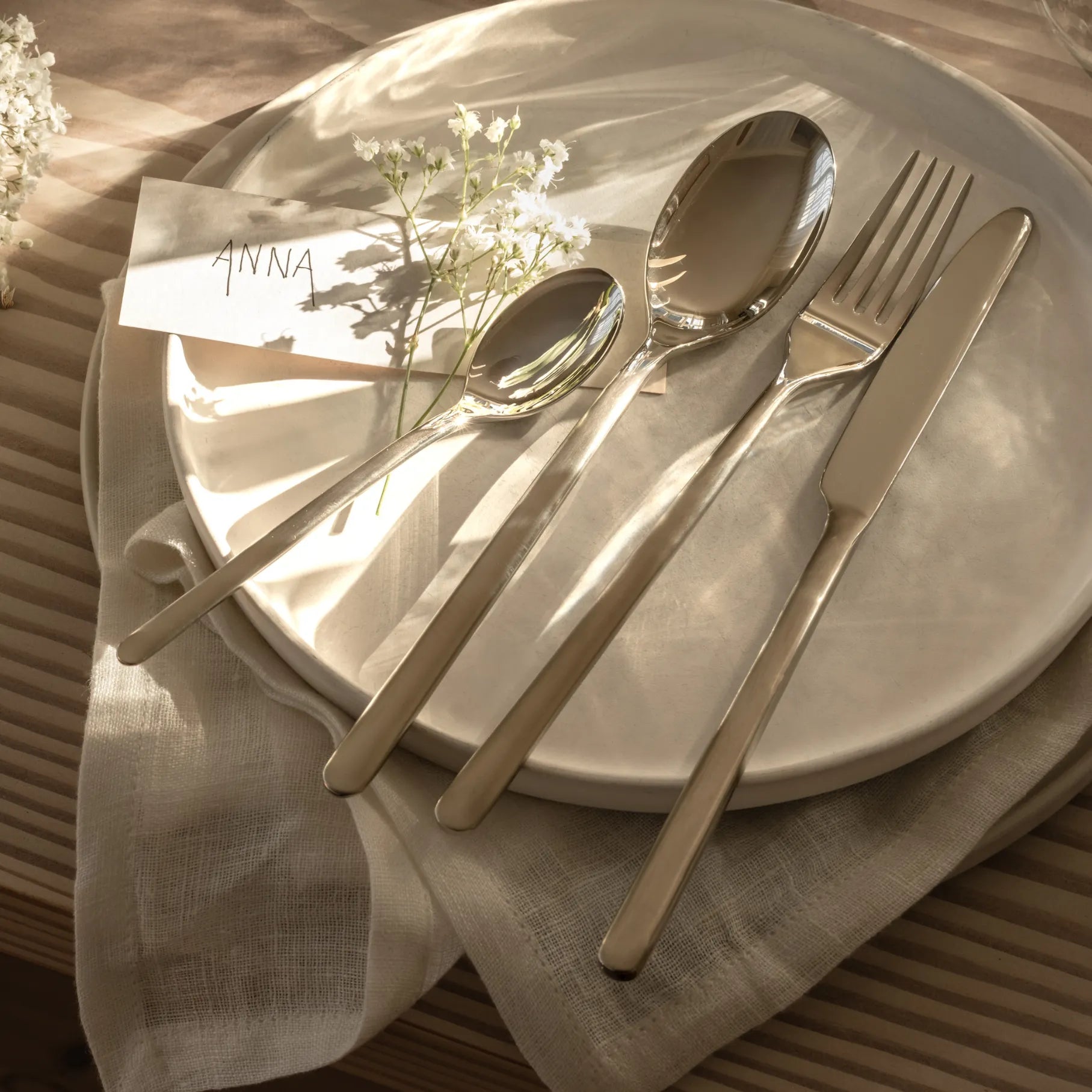 Gense Still Collection cutlery set on a white plate with a name card labeled 'ANNA' on a textured surface.