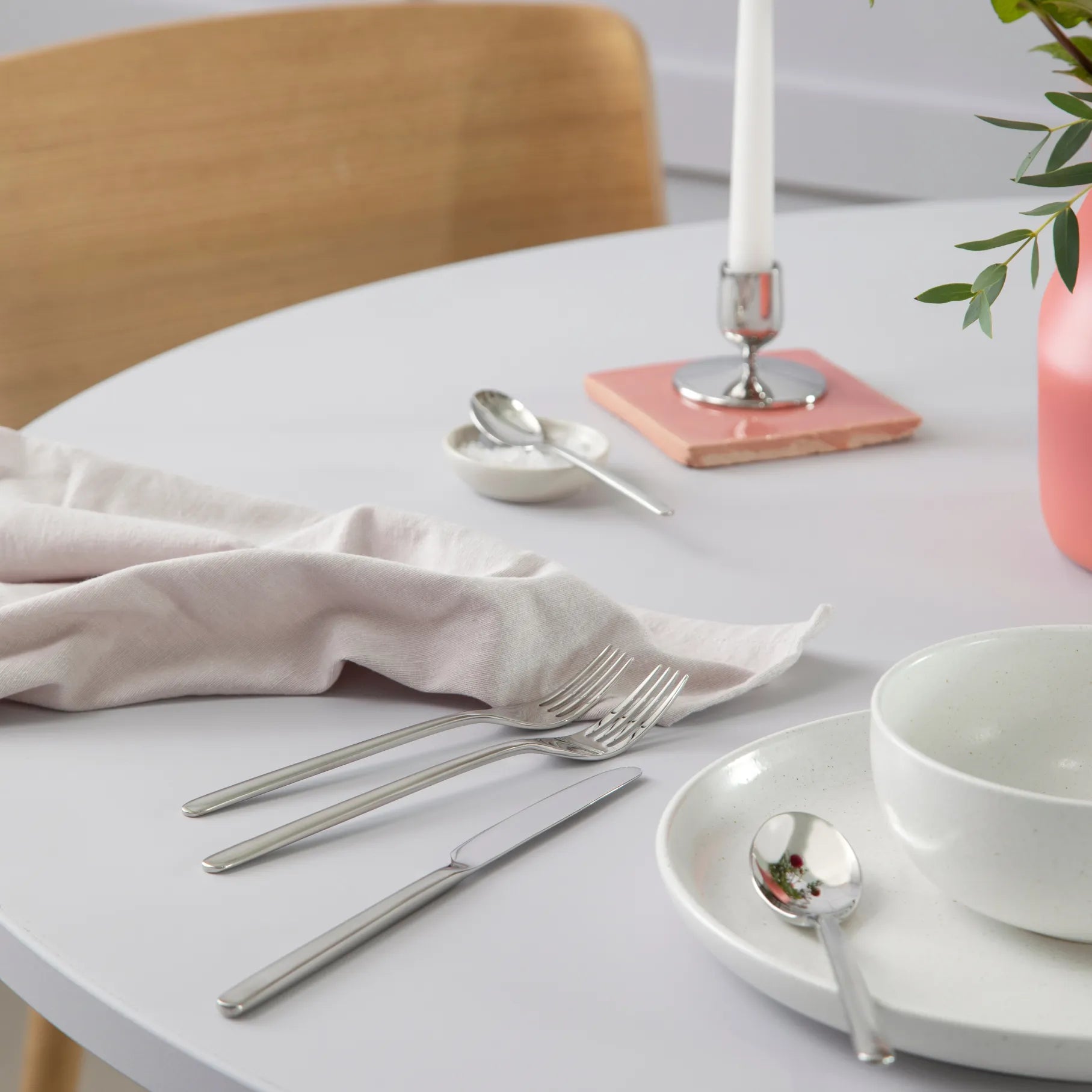 Dining table setting with white plates, Gense Still Collection cutlery, and a pink napkin on a light wooden chair.